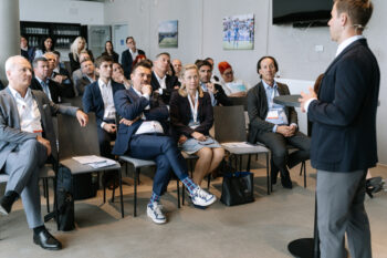 Menschen in Businesskleidung sitzen im Publikum, ein Mann spricht vor der Gruppe